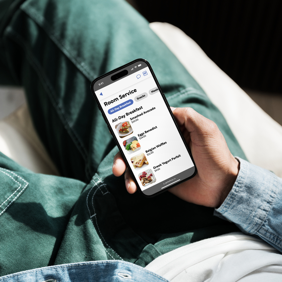 A guest sitting down holding a smartphone in their lap displaying the room service section of the Bellhop guest compendium system. The category “All Day Breakfast” is currently selected, listing several products including “Smashed Avocado”, “Eggs Benedict”, “Belgian Waffles” and a “Greek Yogurt Parfait”, each accompanied by an appetising photo.